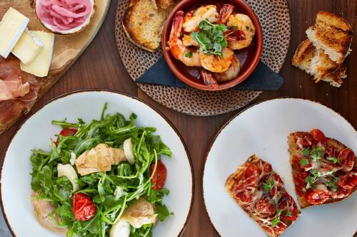 A spread of dishes including shrimp, arugula salad, bruschetta, and toasted bread on wooden table.