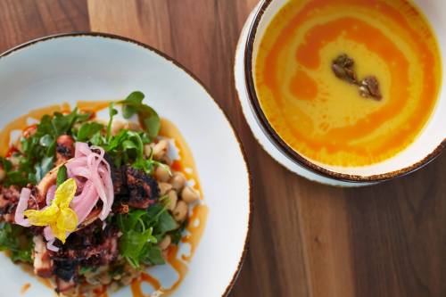 A vibrant dish featuring grilled octopus on a salad with greens and pickled onions, alongside a bowl of yellow soup.