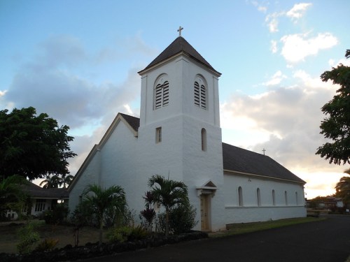 A white church with a tall steeple and cross, surrounded by trees, under a cloudy sky at sunset.