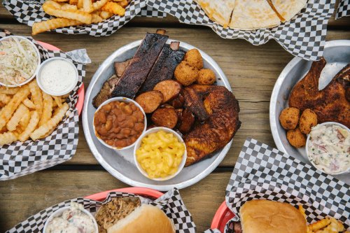 A spread of barbecue dishes including ribs, sausage, chicken, mac and cheese, beans, and fries on checkered paper.