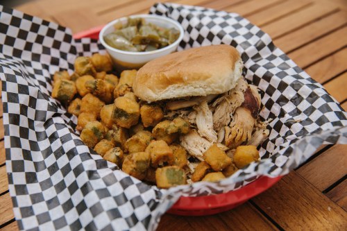 A basket with a pulled chicken sandwich, fried okra, and a side of pickled vegetables on a checkered paper.