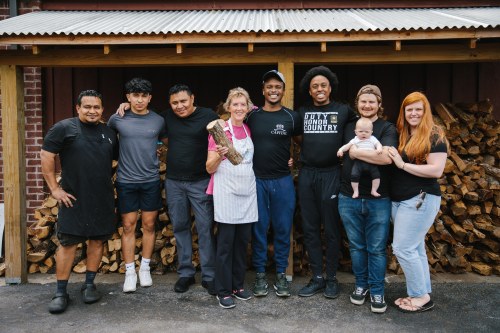 A diverse group of eight people poses together outside a building, smiling, with a stack of firewood in the background.