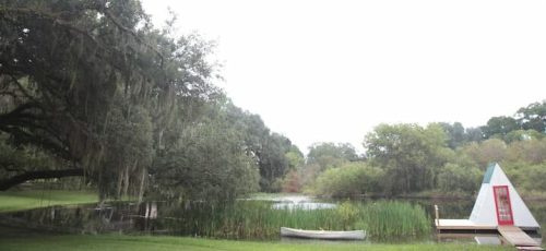 A serene pond surrounded by lush greenery, with a canoe and a small triangular structure on the shore.