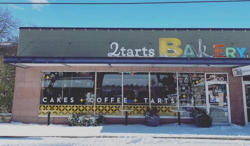 A bakery storefront with large windows displaying "Cakes, Coffee, Tarts" and colorful signage, surrounded by snow.