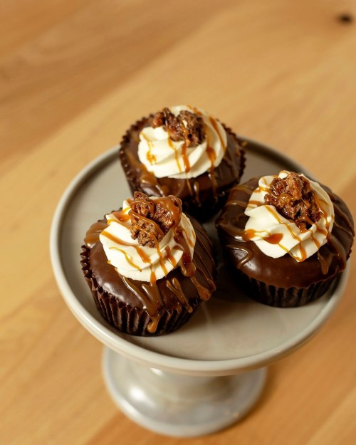 Three chocolate cupcakes with swirls of cream, topped with caramel drizzle and crunchy toppings, on a gray cake stand.