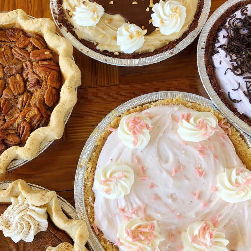 A variety of pies on a wooden table, including pecan, chocolate, and cream pies, all decorated with whipped cream.