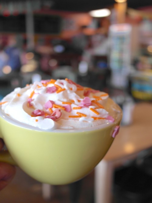 A close-up of a yellow cup filled with whipped cream and colorful sprinkles, set against a blurred café background.