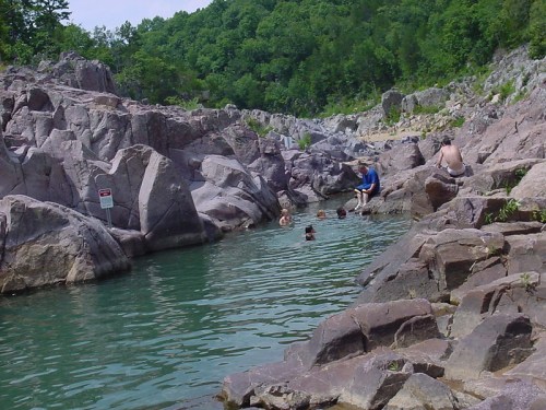 A serene natural pool surrounded by rocky terrain, with people enjoying the water on a sunny day.