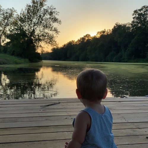 A child sits on a wooden dock, gazing at a serene river and sunset surrounded by trees.