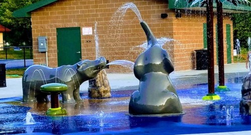A playful splash pad featuring a bear and an elephant sculpture with water jets, surrounded by colorful pavement.