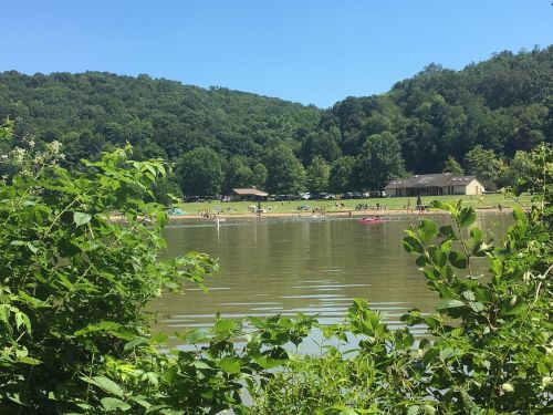 A serene lake scene with people enjoying the beach, surrounded by green hills and trees under a clear blue sky.