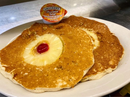 Two pancakes with pineapple rings and a cherry on top, served with a packet of Smucker's breakfast syrup.