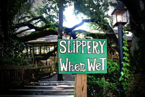 A wooden sign reading "Slippery When Wet" surrounded by trees and a rustic pathway.