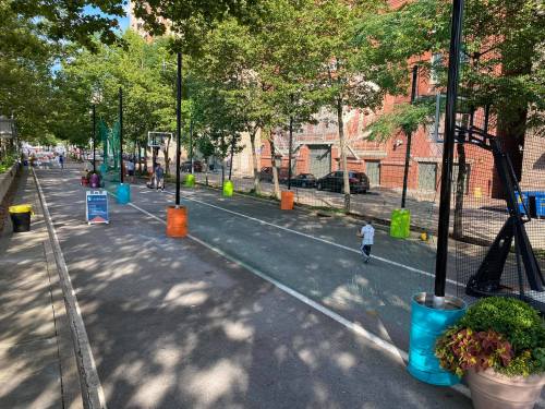 A sunny urban street with a basketball court, colorful barriers, and people enjoying outdoor activities.