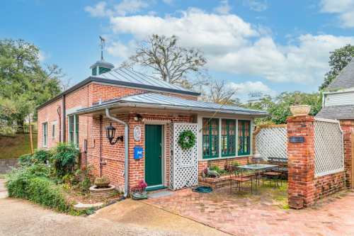 Charming brick cottage with a metal roof, green windows, and a small patio surrounded by greenery and brick pathways.