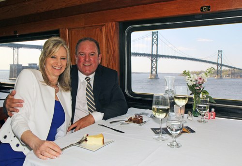 A couple smiles together at a dining table with a view of a bridge and water, enjoying a meal in a train car.