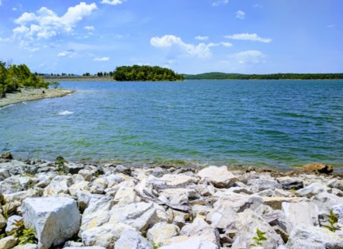 A serene lake scene with rocky shorelines and a small island surrounded by clear blue water under a bright sky.