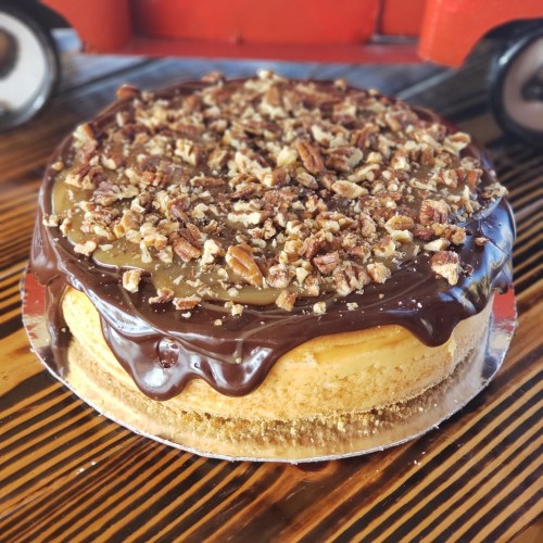 A round cheesecake topped with chocolate ganache, caramel, and chopped pecans on a wooden table.