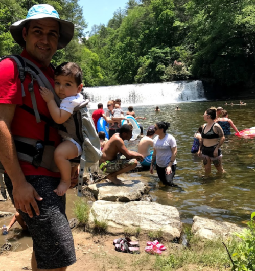 A man holds a baby on his back while people swim and play near a waterfall in a lush, green setting.