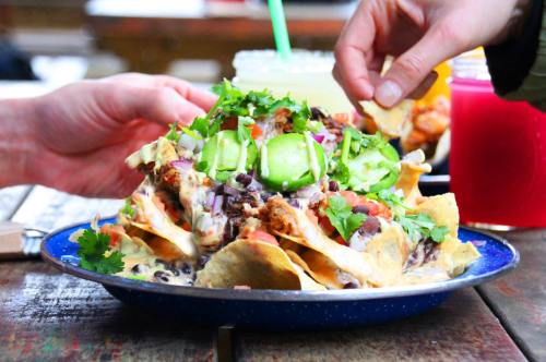 A colorful plate of nachos topped with cheese, jalapeños, tomatoes, onions, and cilantro, with hands reaching in to grab some.