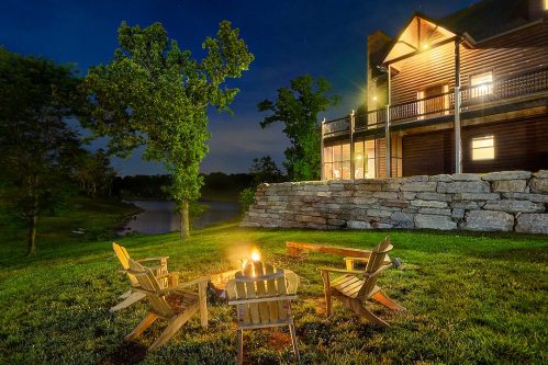 A cozy campfire surrounded by wooden chairs, with a log cabin and serene lake under a starry night sky.