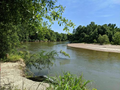 A serene river bends through lush greenery under a clear blue sky, with sandy banks and trees lining the water's edge.