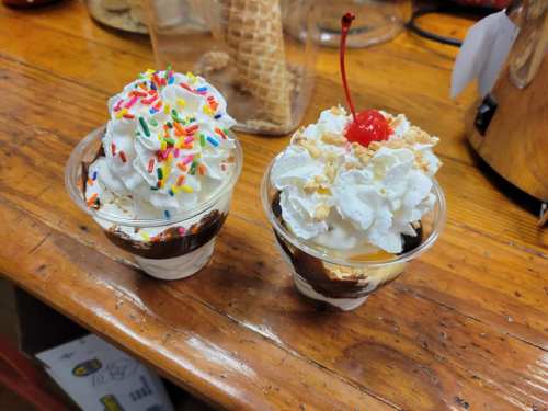 Two dessert cups with whipped cream, one topped with colorful sprinkles and the other with a cherry and crushed nuts.