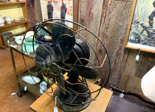 A vintage metal fan with a black finish, displayed on a wooden table in an antique shop.