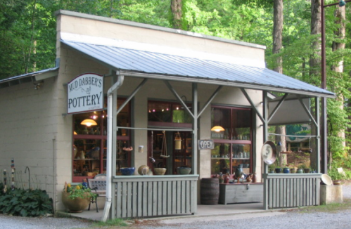 A charming pottery shop with a sign reading "Wild Dabber's Pottery," surrounded by trees and a gravel path.
