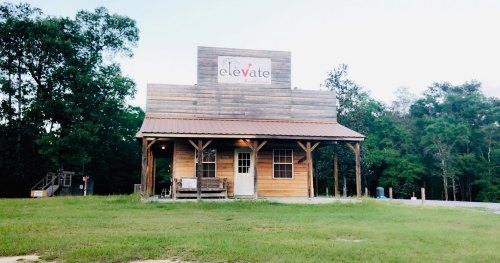 A rustic wooden building with a sign reading "elevate" in a grassy area surrounded by trees.