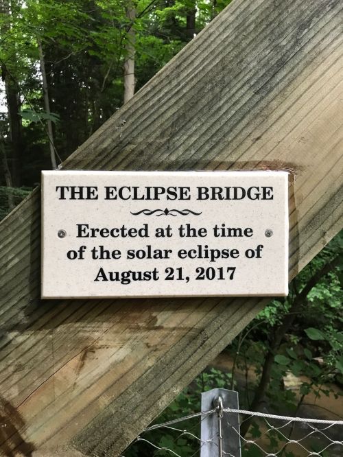 Sign on a wooden post reading "The Eclipse Bridge" with details about its erection during the solar eclipse of August 21, 2017.