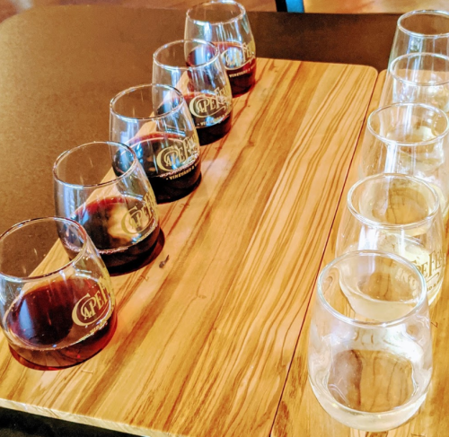 A wooden tasting board with several glasses, some filled with dark liquid and others empty, arranged in two rows.