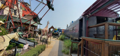 A whimsical amusement area featuring a ferris wheel, giraffe sculptures, and a vintage train alongside a pathway.