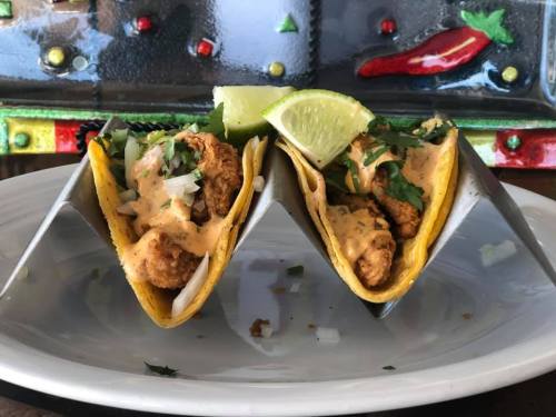 Two tacos filled with fried chicken, topped with sauce, onions, and cilantro, served with lime wedges on a white plate.
