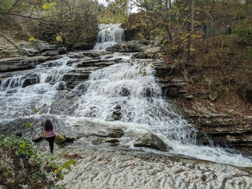 A person stands near a cascading waterfall surrounded by rocky terrain and autumn foliage.