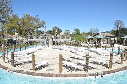 A sunny pool area with lounge chairs, sandy spots, and a wooden walkway surrounded by trees and greenery.
