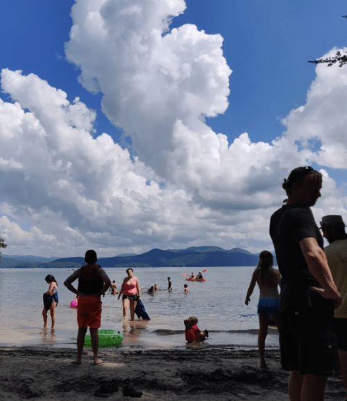 A beach scene with people swimming and playing in the water, surrounded by clouds and mountains in the background.