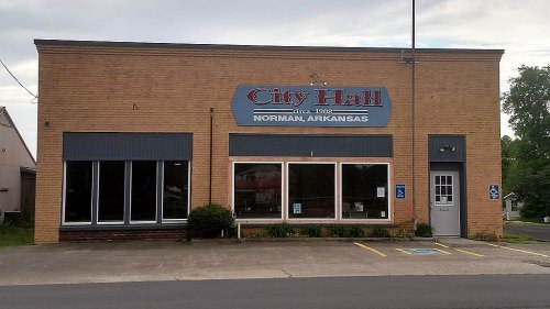Exterior of City Hall building in Norman, Arkansas, featuring large windows and a sign with the town's name and establishment year.