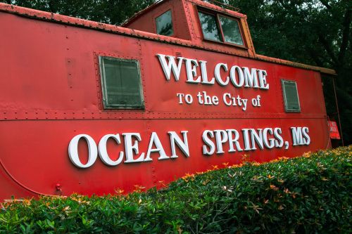 Red train caboose with a sign reading "Welcome to the City of Ocean Springs, MS," surrounded by greenery.