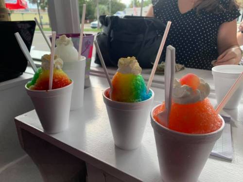 Four colorful snow cones in white cups, topped with whipped cream and straws, on a table.