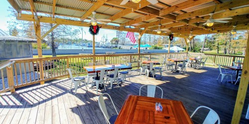 Outdoor dining area with wooden tables, fans, and festive decorations, overlooking a marina and trees.