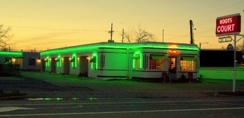 A retro-style motel with green neon lights glowing at dusk, featuring a welcoming entrance.
