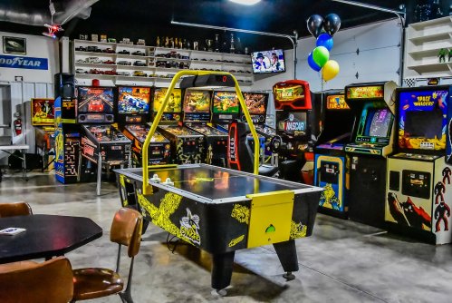 A vibrant arcade featuring various classic pinball and video games, with an air hockey table in the foreground.