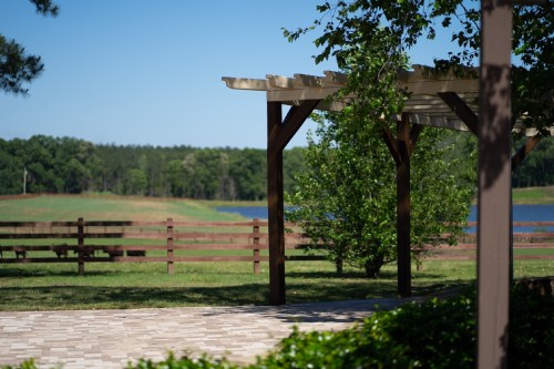A serene outdoor scene featuring a pergola, green grass, a lake, and a wooden fence in the background.