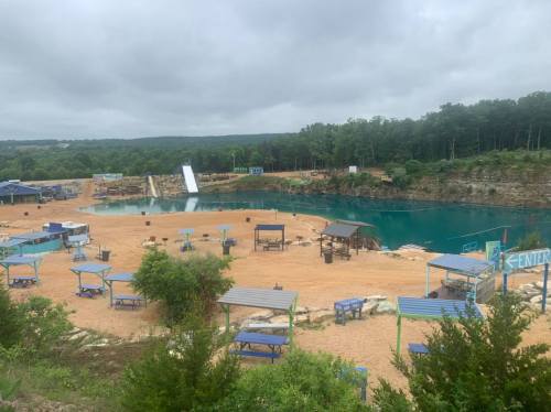 A scenic view of a sandy area with blue water, diving board, and seating, surrounded by trees under a cloudy sky.