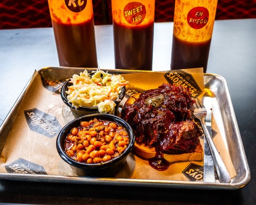 A metal tray with barbecue brisket, coleslaw, baked beans, and three bottles of sauce in the background.