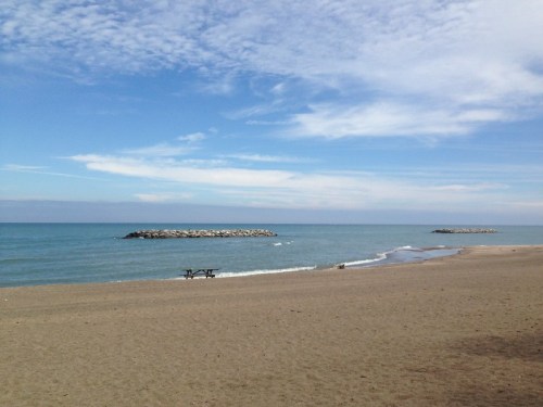 A serene beach scene with calm waters, a sandy shore, and a distant rocky jetty under a blue sky with scattered clouds.
