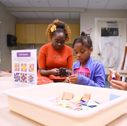 A woman and a girl work on a craft project with colorful paper squares in a bright classroom setting.