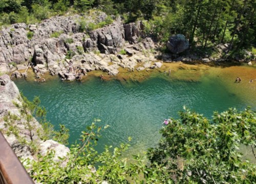 A scenic view of a clear blue swimming hole surrounded by rocky cliffs and lush greenery. People are enjoying the water.