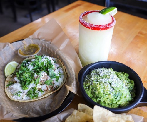 A plate with a taco topped with cilantro and cheese, a bowl of guacamole, tortilla chips, and a frozen margarita.